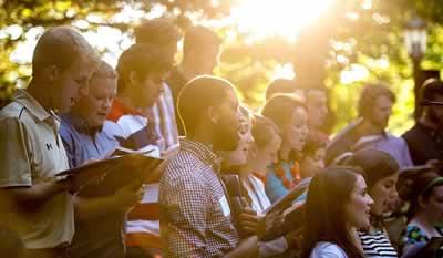 A group of students singing while sunshine pours through the trees above them